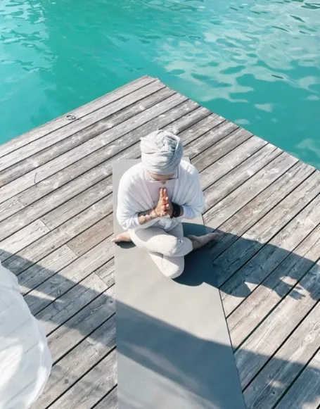 Frau meditiert im Schneidersitz auf einer Yogamatte auf einem Holzsteg über türkisem Wasser