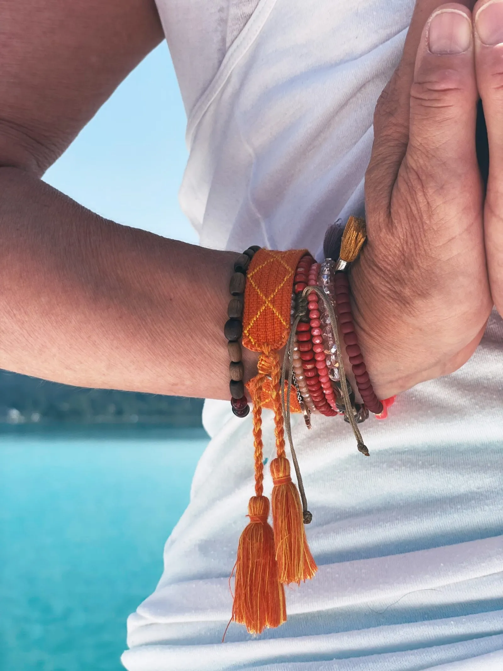 Aneinandergelegte Hände mit bunten Armbändern und Quasten vor dem See
